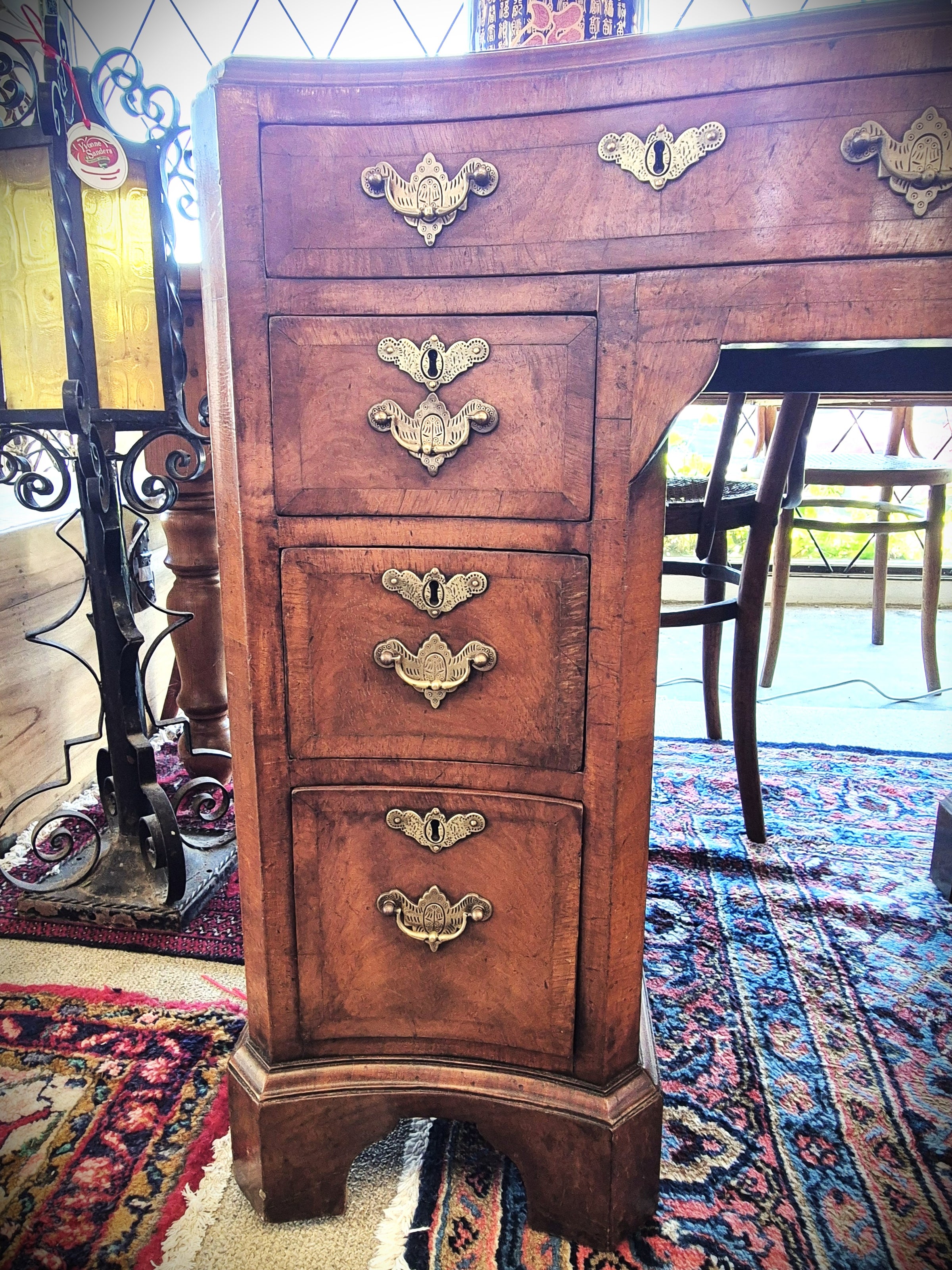 An antique wooden writing desk with four drawers and ornate brass handles, set on a patterned rug in a room.